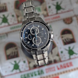 May include: A stainless steel Citizen Eco-Drive watch with a dark blue face and silver accents. The watch features multiple subdials and a tachymeter bezel. The watch band is made of stainless steel.