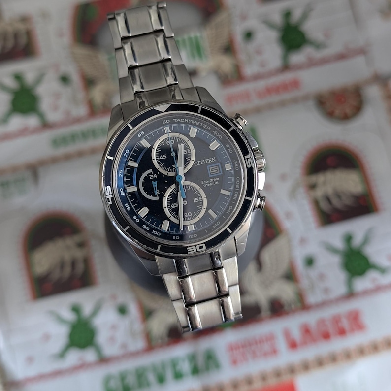 May include: A stainless steel Citizen Eco-Drive watch with a dark blue face and silver accents. The watch features multiple subdials and a tachymeter bezel. The watch band is made of stainless steel.