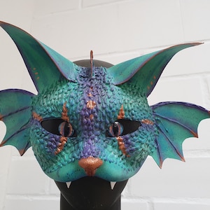 May include: A fantasy cat mask with teal, purple, and copper accents. The mask features large, pointed ears, wing-like structures, and a textured surface. The eyes are dark, and the nose is copper-colored. The mask has sharp teeth.