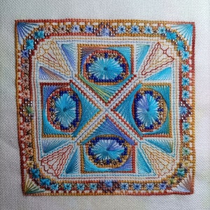 Op de afbeelding: Een vierkant borduurwerk met een geometrisch patroon in tinten blauw, oranje en bruin. Het ontwerp heeft een centraal vierkant met vier kleinere vierkanten eromheen, elk met een uniek patroon.
