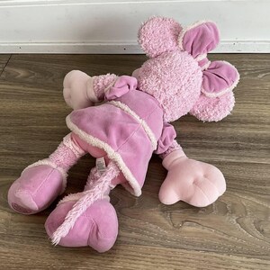 Peut inclure: Une peluche rose en forme de souris avec de grandes oreilles, v&ecirc;tue d'un manteau rose avec des bordures blanches. Le jouet a une longue queue tress&eacute;e et de grands pieds arrondis. Le jouet est pos&eacute; sur une surface en bois.