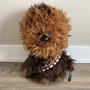 Op de afbeelding: Een Chewbacca pluche pop met bruin en bruin haar. De pop heeft een zwarte neus, blauwe ogen en een bruine en witte bandolier. Een Star Wars-label is bevestigd. De pop staat op een houten vloer.