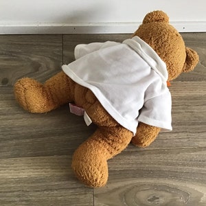 May include: A brown teddy bear wearing a white shirt with a brown bow tie. The bear is lying on a wooden floor.