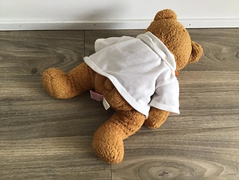 May include: A brown teddy bear wearing a white shirt with a brown bow tie. The bear is lying on a wooden floor.