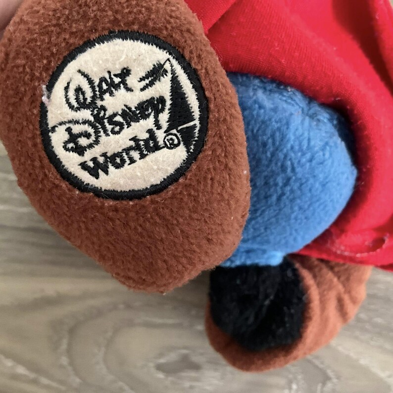 May include: Close-up of a plush toy featuring brown, blue, and red fabric. A circular patch with "Walt Disney World" text and logo is sewn onto the brown fabric. The toy's design is partially visible.