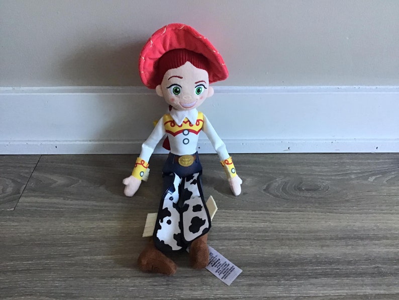 May include: A plush toy of Jessie from the Toy Story franchise. She is wearing a red hat, a white shirt with yellow accents, and blue jeans with a cow print. She is sitting on a wooden floor.