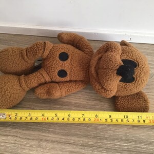 Puede incluir: Un juguete de peluche marr&oacute;n con forma de perro de dibujos animados. El juguete tiene ojos negros y una nariz negra. El juguete est&aacute; tumbado sobre una superficie de madera.