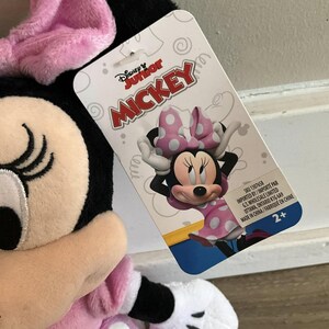 May include: Pink and black plush Minnie Mouse toy with a pink bow and white polka dots. The toy is holding a pink and white polka dot bow. The toy is 2+ years old. The packaging says "Disney Junior Mickey".