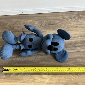 K&ouml;nnte beinhalten: Ein blaues Pl&uuml;schtier in Form einer Zeichentrickmaus, das auf einer Holzoberfl&auml;che liegt. Das Spielzeug hat schwarze Knopfaugen und eine schwarze Nase. Ein gelbes Ma&szlig;band liegt neben dem Spielzeug.