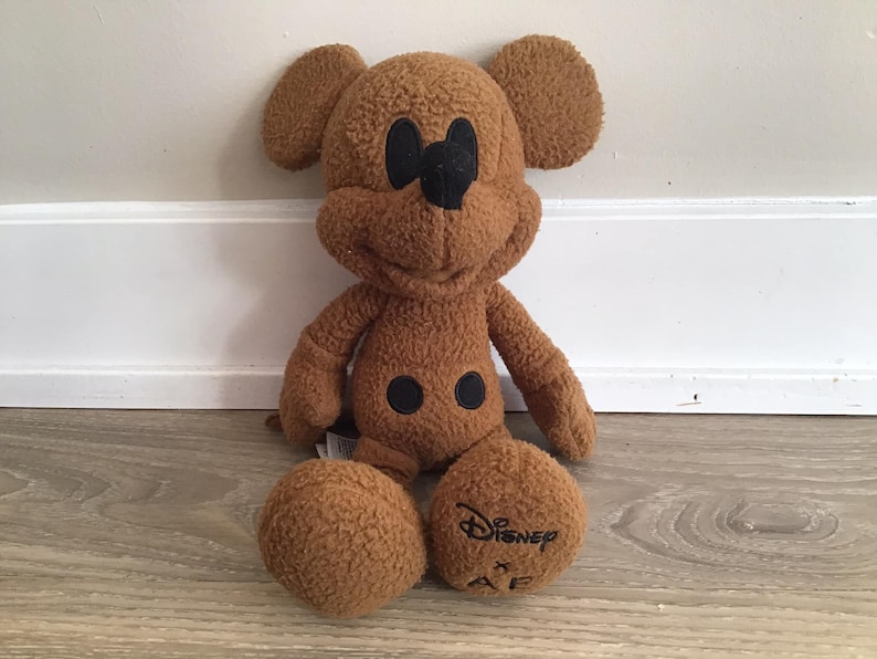 Puede incluir: Un juguete de peluche Mickey Mouse marr&oacute;n con ojos y botones negros. El juguete est&aacute; sentado en un suelo de madera y tiene el texto "Disney x AF" en su pie.