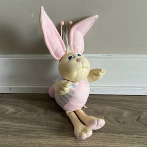 May include: A plush Easter bunny doll with a cream-coloured face and body, large pink ears, and blue eyes. The bunny wears a pink dress with a striped pattern and has long, floppy legs. The doll is sitting on a wooden floor.