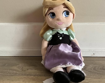 May include: A plush doll of a princess with blonde hair, blue eyes, and a pink mouth. The doll wears a black bodice, a light green top, and a purple skirt with white lace trim. Black shoes complete the outfit.