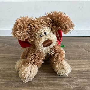 May include: A brown and beige plush dog toy with a red bow around its neck. The toy has a stitched brown nose and black eyes.