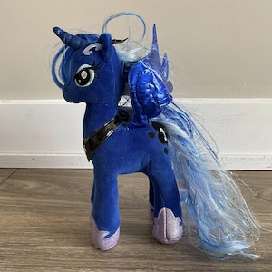 Puede incluir: Un peluche de la Princesa Luna de My Little Pony. El pony es principalmente azul oscuro con una larga melena y cola fluidas en tonos azules y blancos. Tiene alas iridiscentes y pezuñas lavanda.