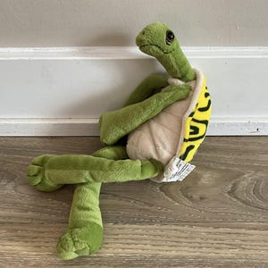May include: A plush green turtle toy with a cream-colored belly and a yellow shell with black markings. The turtle has a long neck, and its legs are extended. The toy is sitting on a wooden surface.
