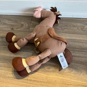 Puede incluir: Caballo de peluche marr&oacute;n con una silla de montar y una etiqueta de Disney. El caballo tiene la boca muy abierta y est&aacute; tumbado de lado.