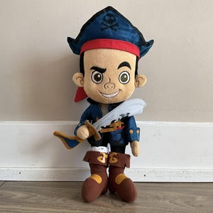 May include: A plush pirate doll wearing a blue pirate hat with a skull and crossbones, a red headband, and a blue jacket. The doll is holding a toy sword and has brown boots. The doll is a cartoon character.