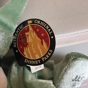 May include: A green plush toy with a black and white face. The toy has a tag that says "Authentic Original Disney Parks" with a picture of Cinderella's castle and a price tag of $10.95.