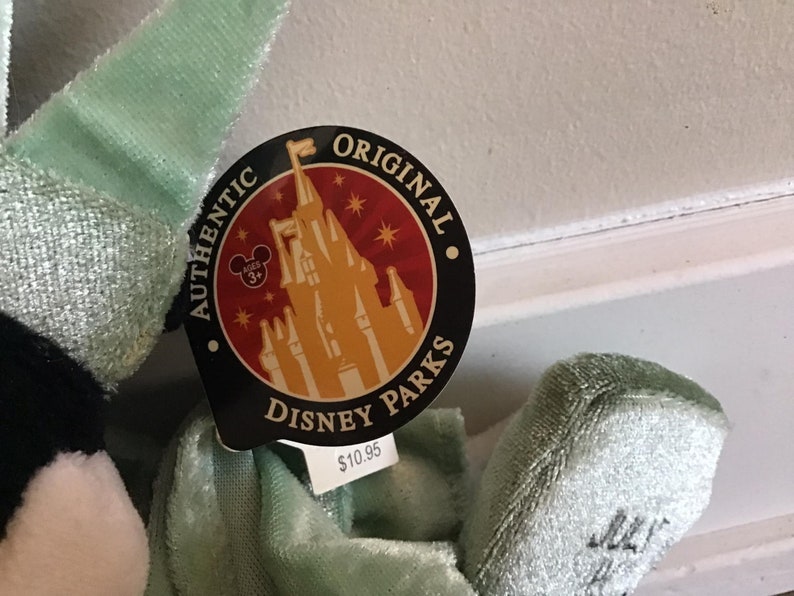 May include: A green plush toy with a black and white face. The toy has a tag that says "Authentic Original Disney Parks" with a picture of Cinderella's castle and a price tag of $10.95.