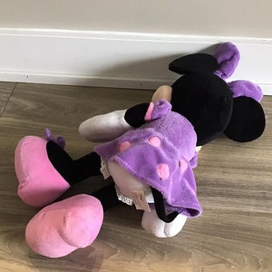 May include: A purple and black plush toy with pink shoes. The toy is lying on a wooden floor. The toy has a purple dress with white polka dots and pink accents.