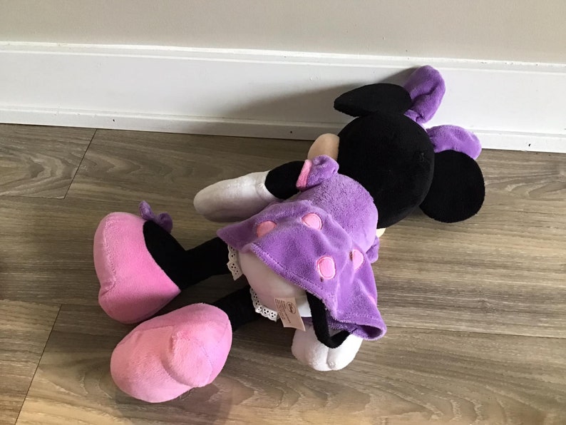 May include: A purple and black plush toy with pink shoes. The toy is lying on a wooden floor. The toy has a purple dress with white polka dots and pink accents.
