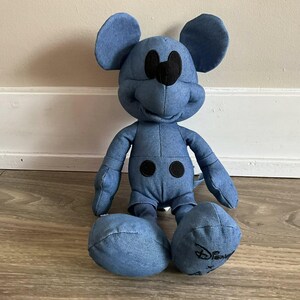 K&ouml;nnte beinhalten: Ein Mickey Mouse Pl&uuml;schtier im Jeans-Stil. Das Pl&uuml;schtier hat ein blaues Jeans-Aussehen mit schwarzen Akzenten f&uuml;r Augen, Nase und Kn&ouml;pfe. Das Disney-Logo ist auf einem Fu&szlig; gestickt. Das Spielzeug sitzt auf einem Holzboden.