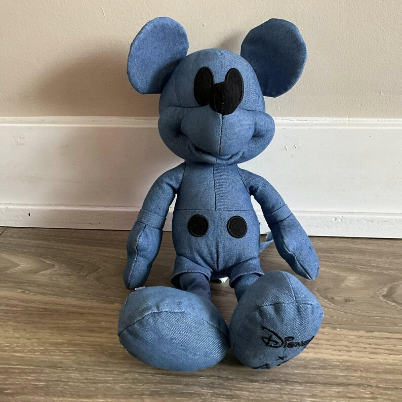 K&ouml;nnte beinhalten: Ein Mickey Mouse Pl&uuml;schtier im Jeans-Stil. Das Pl&uuml;schtier hat ein blaues Jeans-Aussehen mit schwarzen Akzenten f&uuml;r Augen, Nase und Kn&ouml;pfe. Das Disney-Logo ist auf einem Fu&szlig; gestickt. Das Spielzeug sitzt auf einem Holzboden.