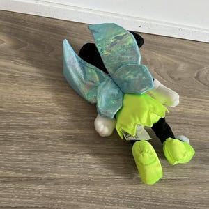 May include: A plush toy with a black head, white hands, and lime green feet. It has iridescent blue fairy wings and a lime green skirt. The toy is on a wooden surface.
