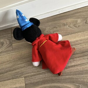 May include: A plush toy of a mouse character wearing a blue wizard hat with white stars and a red robe with yellow trim. The mouse has black ears and a white face. The toy is lying on a wooden floor.
