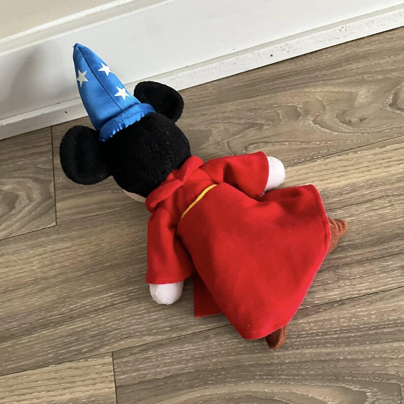 May include: A plush toy of a mouse character wearing a blue wizard hat with white stars and a red robe with yellow trim. The mouse has black ears and a white face. The toy is lying on a wooden floor.