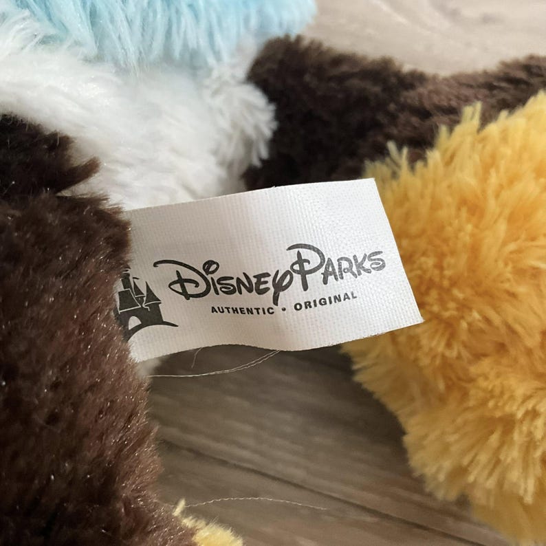 May include: Close-up of a Disney Parks tag with the text "AUTHENTIC &bull; ORIGINAL". The tag is white with the Disney Parks logo and is attached to a brown, yellow, and blue plush toy.