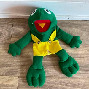 May include: A green frog hand puppet featuring a bright red mouth with a yellow tongue, large white eyes with black pupils, and yellow fabric overalls with two brown buttons attached to the front straps, constructed from soft plush material.