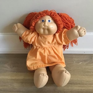 May include: A vintage Cabbage Patch Kid doll with orange hair, blue eyes, and a peach-colored dress. The doll is sitting on a wooden floor.