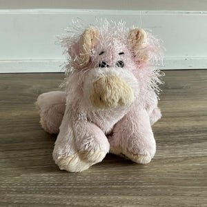 Puede incluir: Un juguete de peluche de cerdo rosa con una melena desgreñada y orejas caídas. El cerdo tiene un hocico y patas de color beige claro y ojos negros pequeños. El juguete está sobre una superficie de madera.