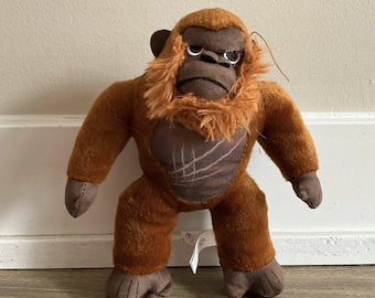 Puede incluir: Un peluche de gorila con pelaje marrón, una melena naranja desgreñada y una expresión severa. El gorila tiene brazos, piernas y orejas de color marrón oscuro. El pecho presenta un parche marrón más oscuro con un diseño cosido. El juguete está de pie.