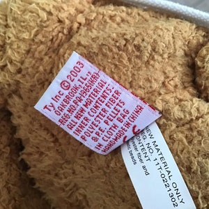 May include: Close-up of a brown plush teddy bear with a white tag attached. The tag reads "Ty Inc &copy;2003 Oakbrook, IL., Reg. No. PA-1965(KR) All New Material", and "New Material Only Reg. No. 11T-0221302 Content: Polyester fiber and plastic beads".