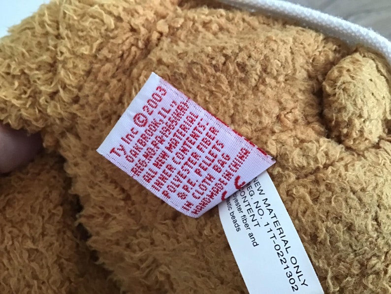 May include: Close-up of a brown plush teddy bear with a white tag attached. The tag reads "Ty Inc &copy;2003 Oakbrook, IL., Reg. No. PA-1965(KR) All New Material", and "New Material Only Reg. No. 11T-0221302 Content: Polyester fiber and plastic beads".