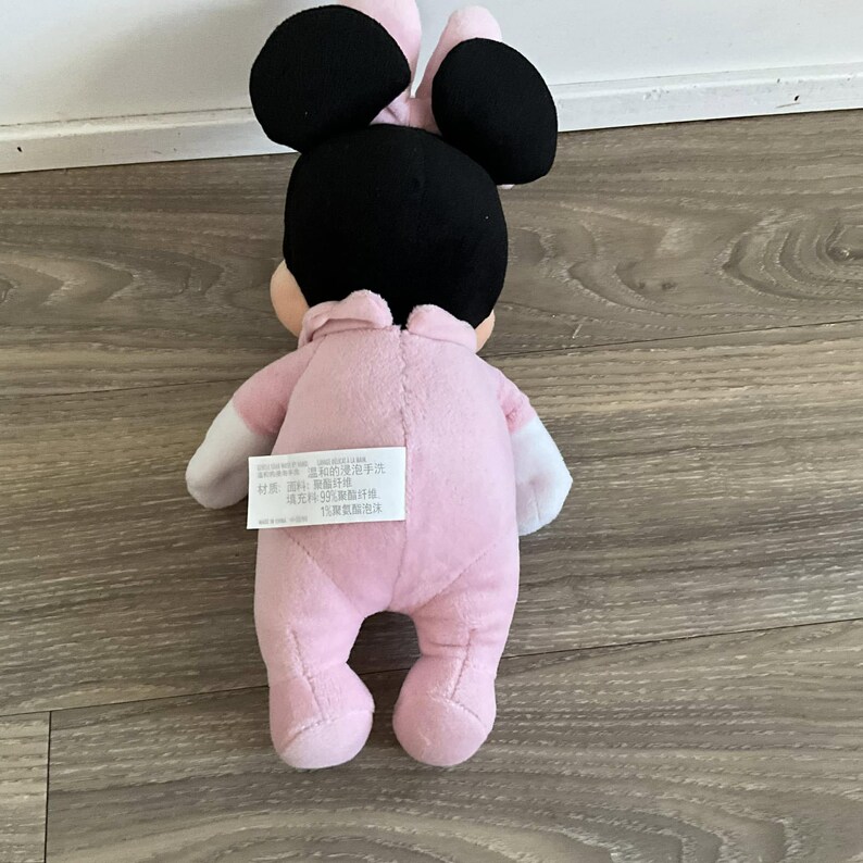 Pu&ograve; includere: Una bambola di peluche di Minnie Mouse, vista da dietro, che indossa una tutina rosa con maniche bianche. La bambola ha orecchie nere e un fiocco rosa. Un'etichetta con testo &egrave; attaccata alla tutina.