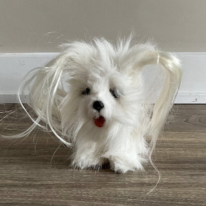 Op de afbeelding: Een witte pluchen speelgoedhond met lang, vloeiend haar en een rode tong. De hond heeft zwarte ogen en een zwarte neus. Het speelgoed staat op een houten oppervlak.