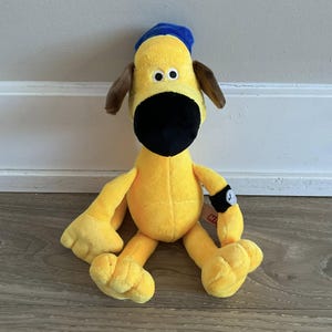 以下が含まれることがあります： 黒い鼻、茶色の耳、青い帽子が特徴の、黄色の犬のぬいぐるみ。おもちゃの腕には、白い時計のデザインが付いた黒いバンドが付いています。木製の表面に置かれています。