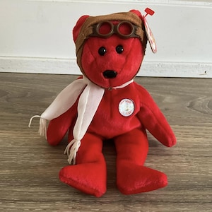 Puede incluir: Un oso de peluche rojo con una gorra de aviador marrón con gafas y una bufanda blanca. El oso tiene un pequeño parche circular que dice "Bear St. Flight". Una etiqueta roja está adherida a la oreja del oso.