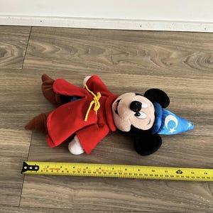 May include: A plush Mickey Mouse toy dressed as a sorcerer, wearing a red robe and a blue hat with a crescent moon and stars. The toy is approximately 12 inches long.