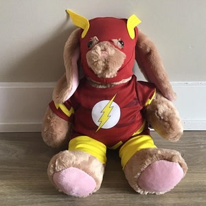Puede incluir: Un conejito de peluche marrón que lleva un disfraz de Flash rojo y amarillo. El disfraz incluye una máscara con un rayo amarillo y un traje rojo y amarillo.