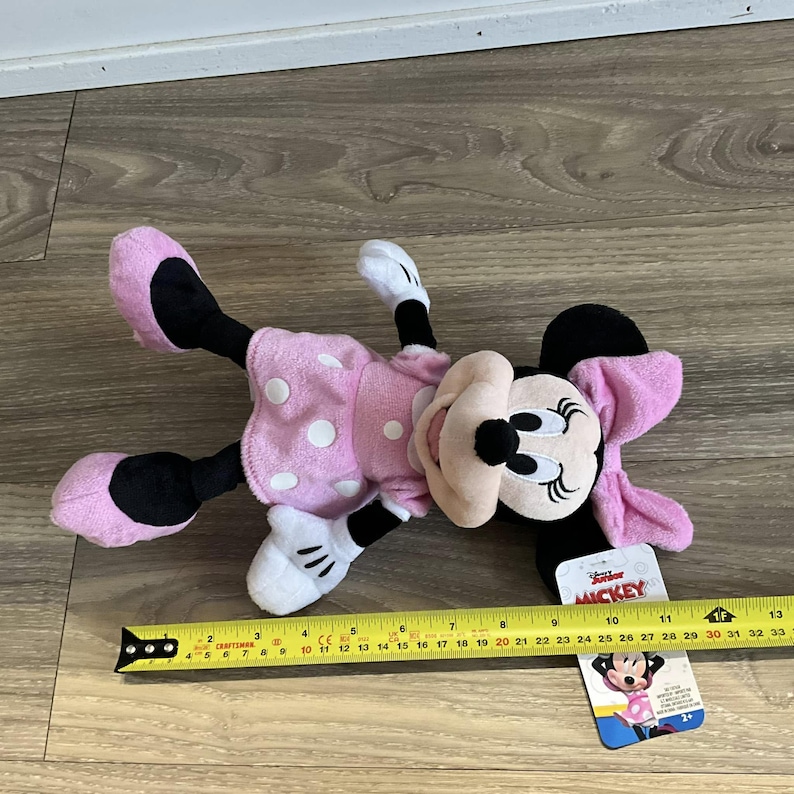 May include: Pink Minnie Mouse plush toy with white polka dots, wearing a pink bow. The toy is lying on a wooden surface with a measuring tape and a tag that says "Mickey".