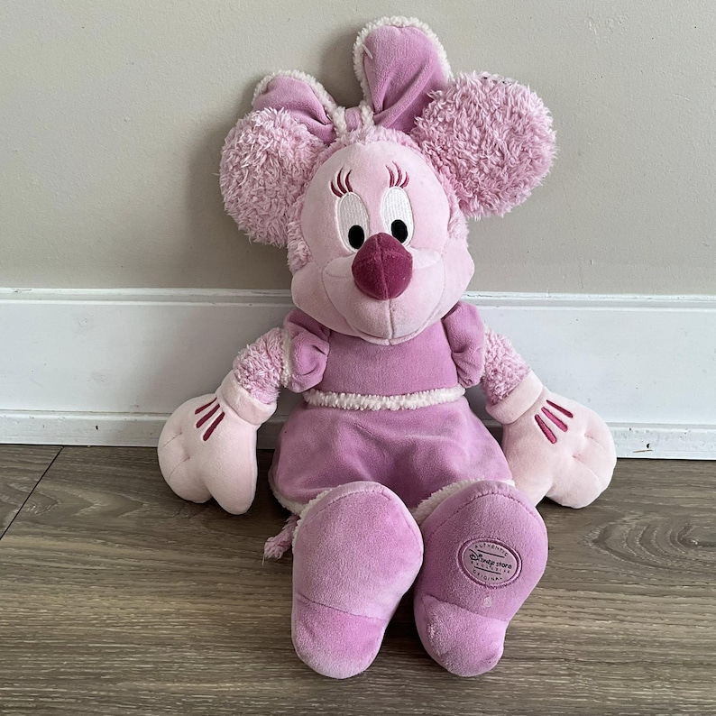 Peut inclure: Une poup&eacute;e Minnie Mouse en peluche rose. La poup&eacute;e a de grandes oreilles duveteuses, un n&oelig;ud et une robe. La poup&eacute;e a un nez rond rose et des yeux blancs. La poup&eacute;e est assise sur un sol en bois.