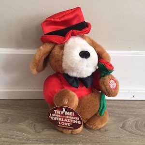 May include: A plush brown dog wearing a red hat and a red jacket with a black bow tie. The dog is holding a red rose and a sign that says "Try Me! I sing and shake! "Everlasting Love" "