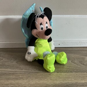 May include: A plush Mickey Mouse doll dressed as a fairy, with black ears, a pink face, and a neon green outfit. The doll has blue iridescent wings and is sitting on a wooden surface.