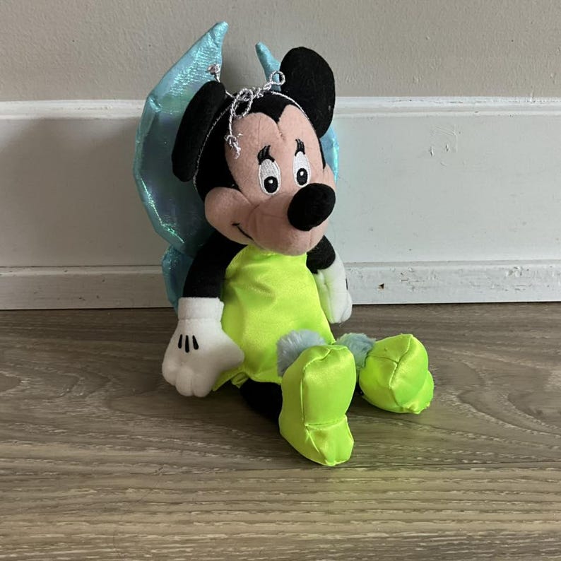 May include: A plush Mickey Mouse doll dressed as a fairy, with black ears, a pink face, and a neon green outfit. The doll has blue iridescent wings and is sitting on a wooden surface.