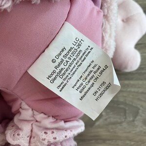 Peut inclure: Gros plan sur une &eacute;tiquette blanche avec du texte noir, attach&eacute;e &agrave; une peluche rose. L'&eacute;tiquette comprend le logo Disney, les informations sur l'entreprise et un code produit. La peluche est rose clair et a des d&eacute;tails en dentelle.