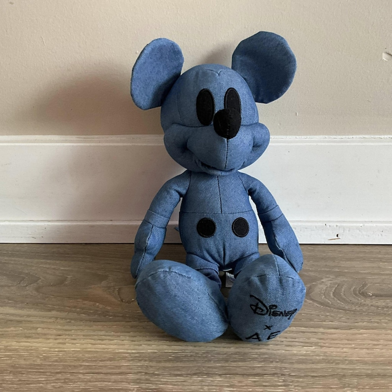 K&ouml;nnte beinhalten: Ein Mickey Mouse-Pl&uuml;schtier aus blauem Denim. Das Spielzeug hat schwarze Augen, eine Nase und zwei schwarze Kreise auf dem Bauch. Das Disney-Logo ist auf einem Fu&szlig; zu sehen. Das Spielzeug sitzt auf einem Holzboden.